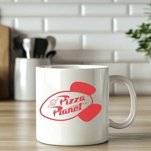 May include: White ceramic mug with a red Pizza Planet logo. The logo features the words "Pizza Planet" in a stylized font, with a planet and rocket ship design. The mug has a curved handle and is set on a wooden surface.