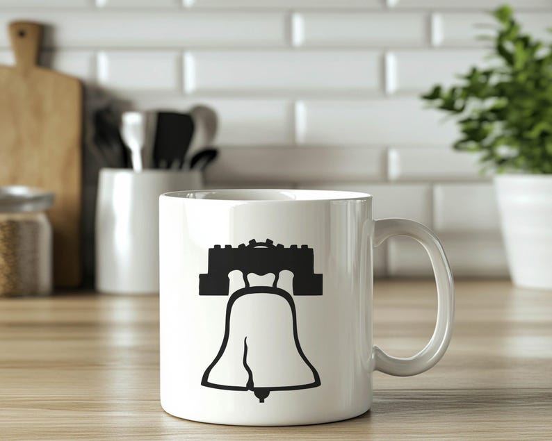 May include: White ceramic mug featuring a black silhouette of the Liberty Bell. The bell is shown with its famous crack.