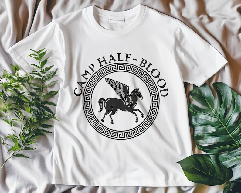 Może przedstawiać: Biała koszulka z czarnym nadrukiem logo Camp Half-Blood. Logo przedstawia skrzydlatego konia w k&oacute;łku z greckim kluczem na obrzeżu i tekstem "Camp Half-Blood".