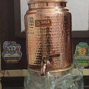 May include: A copper-coloured water dispenser with a hammered texture, a tap, and a lid. The dispenser has a label that reads "SAGA Tableware Collection". The dispenser is partially wrapped in clear plastic.