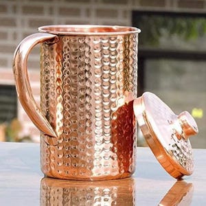 May include: A copper-colored mug with a hammered texture and a matching lid. The mug has a large handle and a cylindrical shape. The lid is round with a small knob. The mug and lid are reflecting the light.