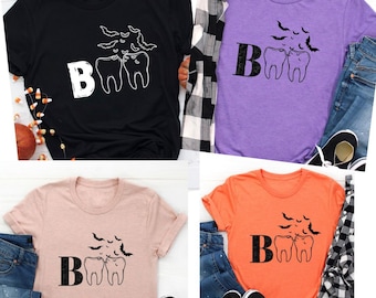 May include: Four t-shirts in black, purple, peach, and orange. Each shirt features the word "Boo" with two cartoon teeth and bats. The shirts are paired with jeans and sneakers, suggesting casual wear.