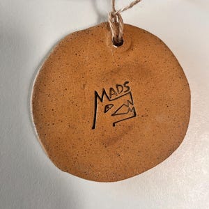 May include: A round, brown ceramic ornament with a textured surface and a small hole at the top for hanging. The word "MADS" is carved into the ornament, along with a stylized design. A natural twine loop is attached.