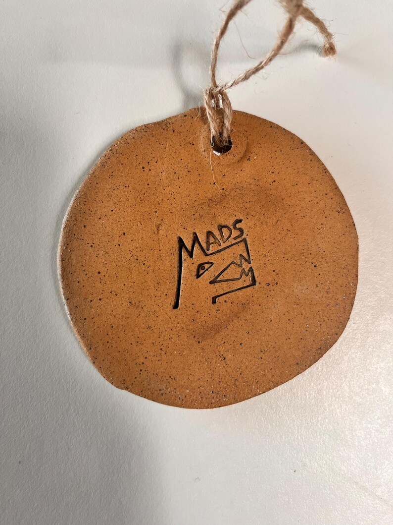 May include: A round, brown ceramic ornament with a textured surface and a small hole at the top for hanging. The word "MADS" is carved into the ornament, along with a stylized design. A natural twine loop is attached.