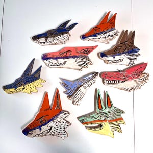 May include: A collection of nine ceramic wolf head sculptures. Each head is uniquely painted with vibrant colors including blue, orange, pink, and brown. The sculptures feature detailed facial expressions and pointed ears, showcasing a whimsical design.