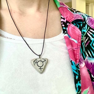 May include: A triangular pendant necklace with a black cord. The pendant features a transgender symbol in black on a light-colored background. The person is wearing a white t-shirt and a colorful floral patterned jacket.