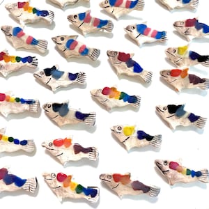 Op de afbeelding: Een verzameling kleine keramische visbeeldjes. Elke vis is wit met kleurrijke accenten, waaronder blauw, rood, geel en paars. De vissen hebben gedetailleerde vinnen en ogen en zijn in een verspreid patroon gerangschikt.