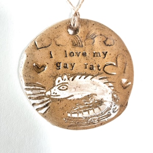 May include: A round, ceramic ornament with the text "I love my gay rat" and a drawing of a rat. The ornament is tan with white accents and a string for hanging. The design includes a rainbow, clouds, and hearts.