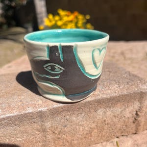 May include: A small ceramic mug with a teal glaze. The mug has a hand-painted design of a black face with a single eye and a heart.