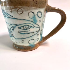 May include: Handmade ceramic mug with a fish design. The mug features a light blue fish illustration on a white background, with blue bubble accents. The rim and handle are a speckled brown. A unique, handcrafted piece for enjoying beverages.