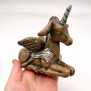 May include: A brown ceramic figurine of a winged unicorn in a reclining position. The creature features a single horn on its forehead, textured wings with white accents, and a glossy finish. The sculpture is crafted with visible carved details.