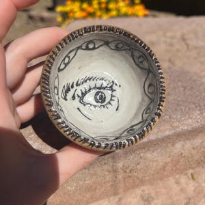 以下が含まれることがあります： 黒い目のデザインが施された小さな白い陶器のボウル。目は黒い瞳と白い虹彩をしています。ボウルは黒い縁取りと縁に黒い目のデザインが施されています。