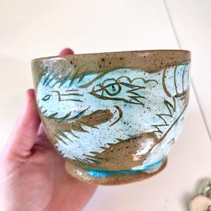 May include: Handmade ceramic bowl with a light blue dragon design. The bowl has a brown rim and base with a speckled texture. The dragon is hand-painted in a light blue color, adding a unique touch to this functional art piece.