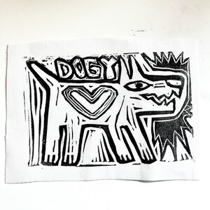 May include: Black and white linocut print of a dog with the word "DOGY" above it. The dog has a heart shape on its chest and a spiky mane.