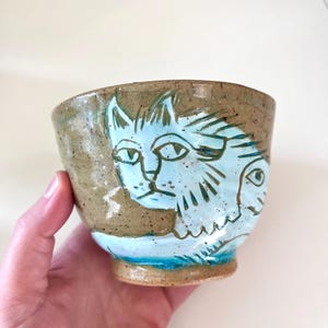 May include: A handmade ceramic bowl with a cat design. The bowl is a speckled brown with a light blue cat face painted on the side. The base of the bowl has a turquoise blue band. The bowl is held in a hand.