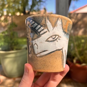 Pode incluir: Uma caneca de cerâmica artesanal com um design de unicórnio extravagante. A caneca apresenta uma base castanha com uma ilustração de unicórnio branco e detalhes em azul escuro e castanho. A caneca é segurada em uma mão, mostrando seu estilo artístico único.