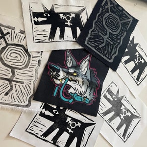 Puede incluir: Un conjunto de parches de tela con varios diseños de perros y lobos. Algunos parches son grabados en linóleo en blanco y negro de un perro estilizado con un símbolo de género. Otros parches tienen un gráfico de cabeza de lobo colorido.