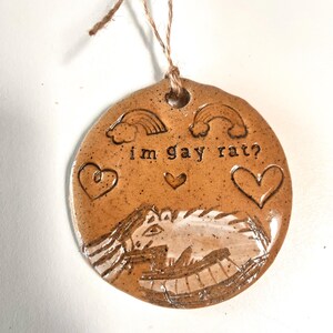 May include: A round, brown ceramic ornament with the text "im gay rat?" and a drawing of a rat. The ornament also features two rainbows, three hearts, and a jute string for hanging.