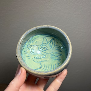 May include: A small ceramic bowl with a light green glaze and a blue wolf design. The wolf's face is outlined in blue and has a detailed design.