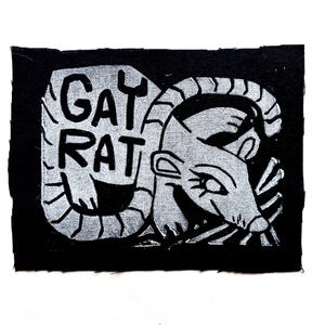 May include: A black patch with a white illustration of a rat and the words "GAY RAT". The rat is stylized with a smiling face and detailed whiskers. The text is in a bold, blocky font.