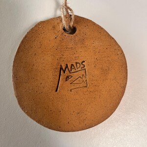May include: A round, brown ceramic ornament with a natural, speckled texture. The ornament has a hole at the top for hanging with a twine string. The word "MADS" is stamped on the surface, along with a small graphic.
