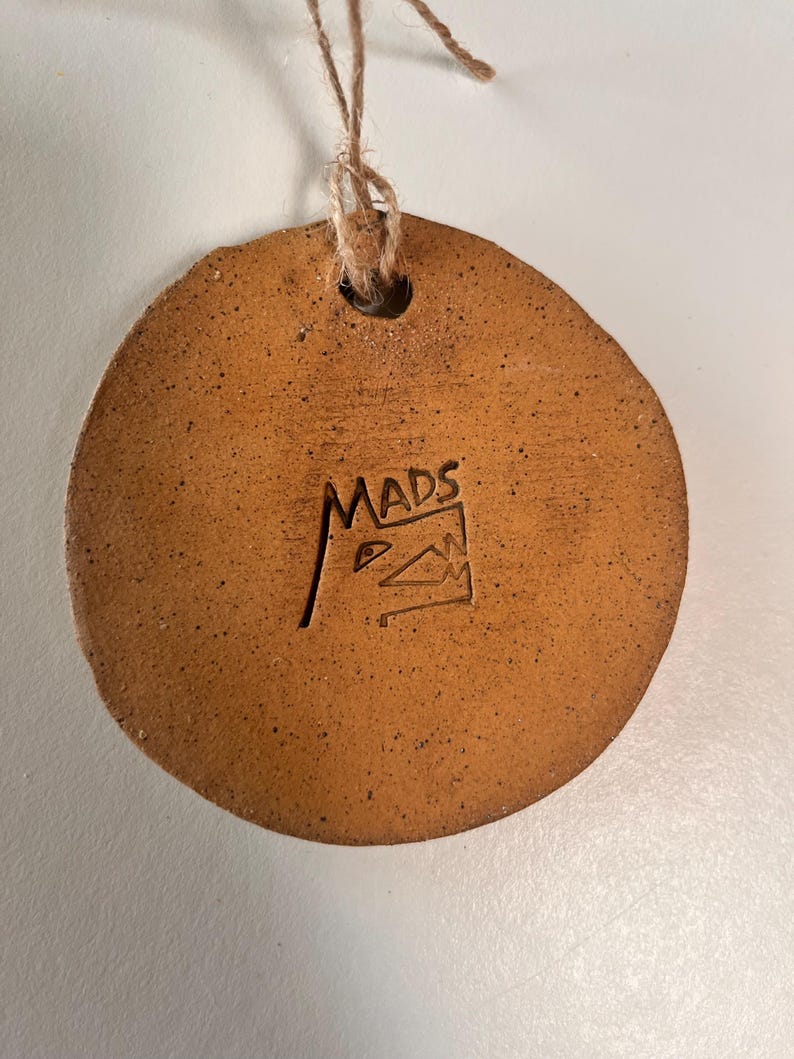 May include: A round, brown ceramic ornament with a natural, speckled texture. The ornament has a hole at the top for hanging with a twine string. The word "MADS" is stamped on the surface, along with a small graphic.