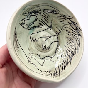 May include: A round, light green ceramic bowl featuring a black line drawing of a wolf-like creature curled in the center. The creature has sharp teeth, pointed ears, and textured fur lines, with the design etched into the glazed surface.