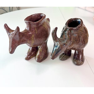 Puede incluir: Dos figuras de rinoceronte de cerámica en tonos marrones y verdes. Los rinocerontes tienen una superficie texturizada y una parte superior ahuecada, lo que sugiere que podrían usarse como pequeños jarrones o piezas decorativas. Las figuras están sobre una superficie blanca.