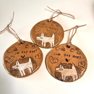 May include: Three handmade ceramic ornaments with a brown glaze. Each ornament features a white dog illustration, hearts, rainbows, and the text "im gay dog?". They are strung with twine for hanging.