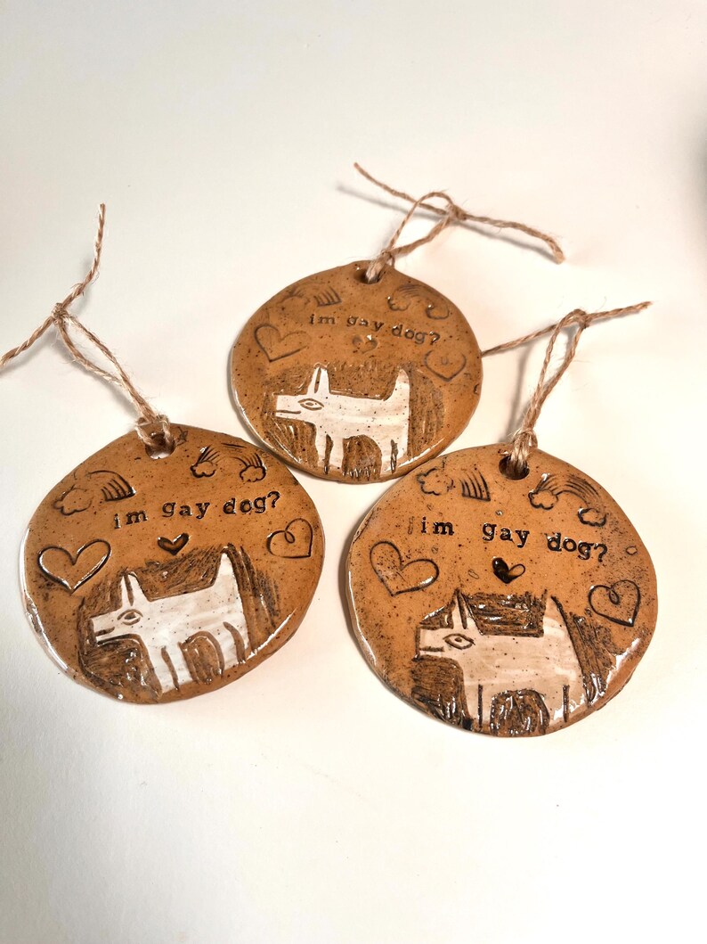 May include: Three handmade ceramic ornaments with a brown glaze. Each ornament features a white dog illustration, hearts, rainbows, and the text "im gay dog?". They are strung with twine for hanging.
