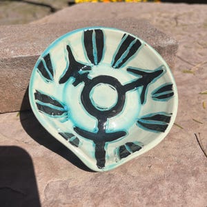 Puede incluir: Un pequeño plato de cerámica blanca con un esmalte turquesa. El plato tiene un diseño en blanco y negro de un símbolo transgénero con hojas estilizadas alrededor.