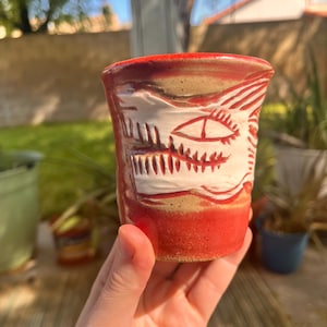 May include: A handmade ceramic cup with a red base and rim, featuring a carved white face design. The cup has a tan middle section and is held in a person's hand. The design includes an eye and teeth.