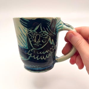 May include: A ceramic mug with a cream-colored base and a dark blue, sgraffito-style carved illustration of a face. The design features etched lines revealing the lighter clay beneath the dark glaze, with a curved handle and a flared rim.