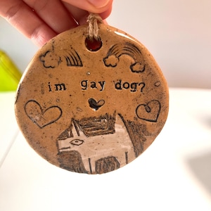 May include: A circular, brown ceramic ornament with the text "im gay dog?" and a dog illustration. The ornament has etched rainbows, hearts, and a hole for hanging with a twine string.