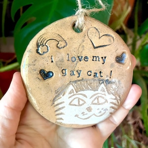 May include: A circular, tan ceramic ornament with the text "i love my gay cat!" and a cat face illustration. The ornament features a rainbow, hearts, and a jute string for hanging. A green plant forms the background.