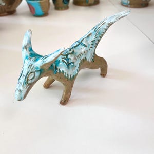Puede incluir: Una escultura de cerámica de un animal estilizado con cuello largo y cuernos curvos. La escultura es principalmente marrón con detalles turquesa y blanco. Se ven otras tazas y tazas de cerámica en el fondo.