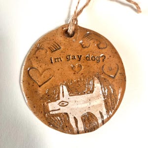May include: A round, brown ceramic ornament with the text "im gay dog?" and a white dog illustration. The ornament has a twine loop for hanging and features heart and rainbow designs.