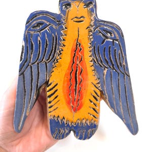 Puede incluir: Una pieza de arte de cerámica con forma de pájaro o ángel estilizado. Las alas y la cabeza son azules con detalles negros, mientras que el cuerpo es amarillo con un diseño central rojo y naranja. La pieza se sostiene en una mano.