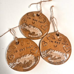 May include: Three brown ceramic ornaments with the text "im gay rat?" and a rat illustration. Each ornament has a hole at the top with a twine string for hanging. The design includes hearts and rainbows.