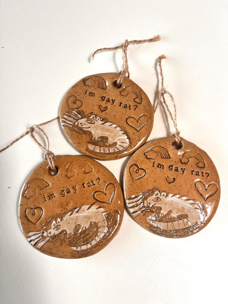 May include: Three brown ceramic ornaments with the text "im gay rat?" and a rat illustration. Each ornament has a hole at the top with a twine string for hanging. The design includes hearts and rainbows.