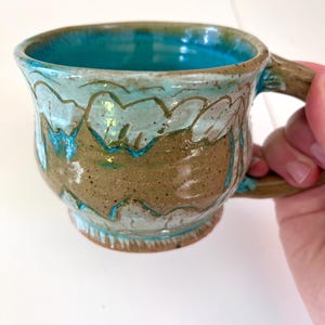 Könnte beinhalten: Handgefertigte Keramik-Tasse mit türkisfarbenem Innenraum und braunem Äußeren. Die Tasse hat ein einzigartiges Design mit hellblauen und braunen Akzenten. Der Griff ist braun.
