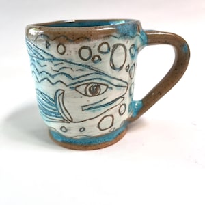 May include: Handmade ceramic mug with a unique design. The mug features a white base with blue and brown accents. The design includes an eye and abstract shapes. The handle and rim are brown, while the interior and base have a blue glaze.