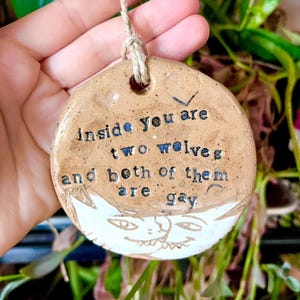 May include: A round, ceramic ornament with the text "inside you are two wolves and both of them are gay." The ornament has a cat face design at the bottom and a jute string for hanging. The text is in black and blue.