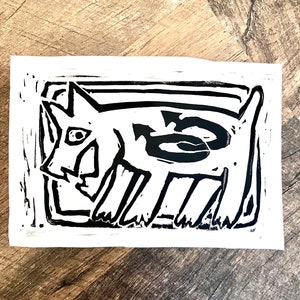 May include: Black and white linocut print of a stylized dog with a large eye. The dog is framed in a black rectangle.