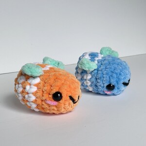 Puede incluir: Dos juguetes de peluche tejidos a crochet con caras sonrientes. Uno es naranja y blanco con orejas verdes, el otro es azul y blanco con orejas verdes.