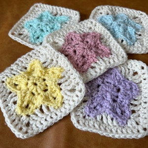 May include: Five crocheted squares with a star design in different colors. The squares are made with white yarn and have a star design in the center. The stars are in shades of light blue, pink, yellow, and purple.