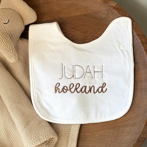 Puede incluir: Babero blanco para bebé con bordado marrón que dice "JUDAH holland".