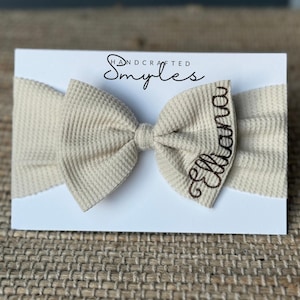 May include: A beige waffle-knit baby headband with a bow and the name "Elliana" embroidered in brown script. The headband is displayed on a white card with the text "Handcrafted Smyles".