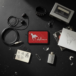 May include: A collection of music-themed items on a black surface. Includes a red and white Deftones patch, a cassette player, headphones, a bracelet, a ring, guitar picks, a lighter, and album art.