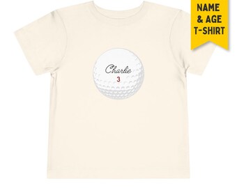 May include: A cream-colored short-sleeve t-shirt featuring a centered graphic of a white golf ball. The golf ball design includes the name Charlie in black cursive script and the number 3 in red. A yellow tag in the corner reads CUSTOM NAME & AGE T-SHIRT.